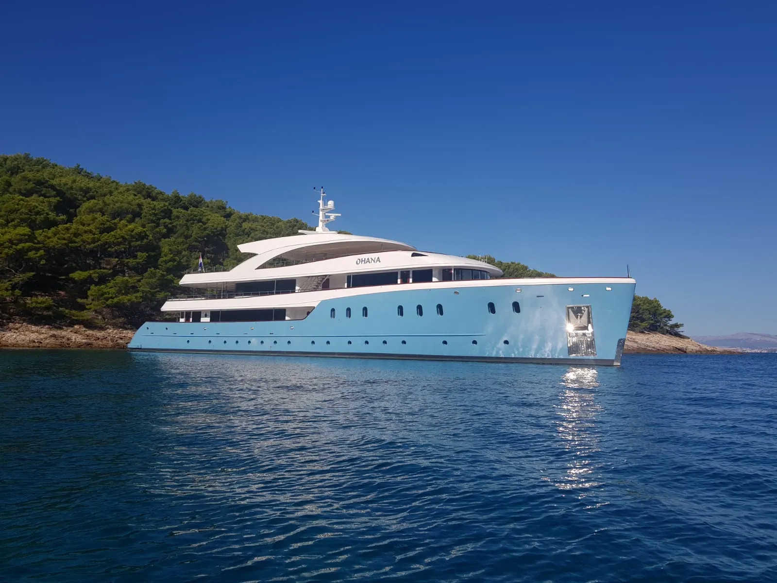 MY Ohana anchored in a calm blue bay with a wooded shore